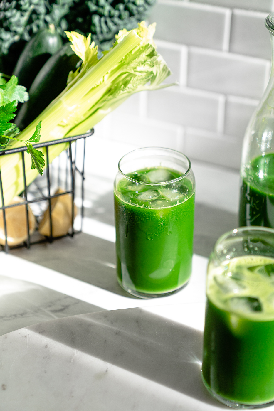 Two glasses of vibrant green juice and basket full of celery, kale, cucumbers, and lemon in the background.