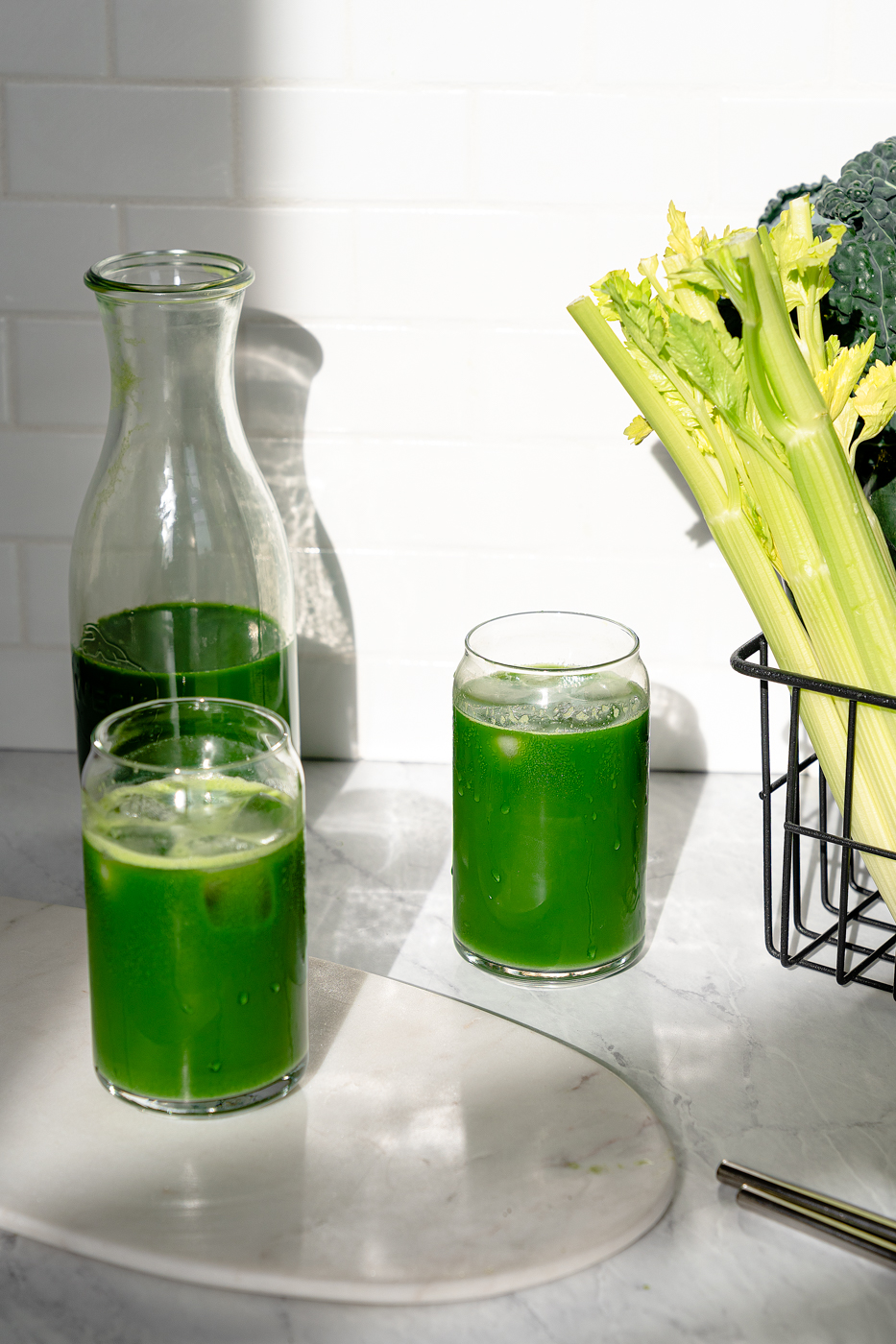 Two glasses of vibrant green juice with a jug of green juice and a basket full of celery, kale, and cucumbers in the background.