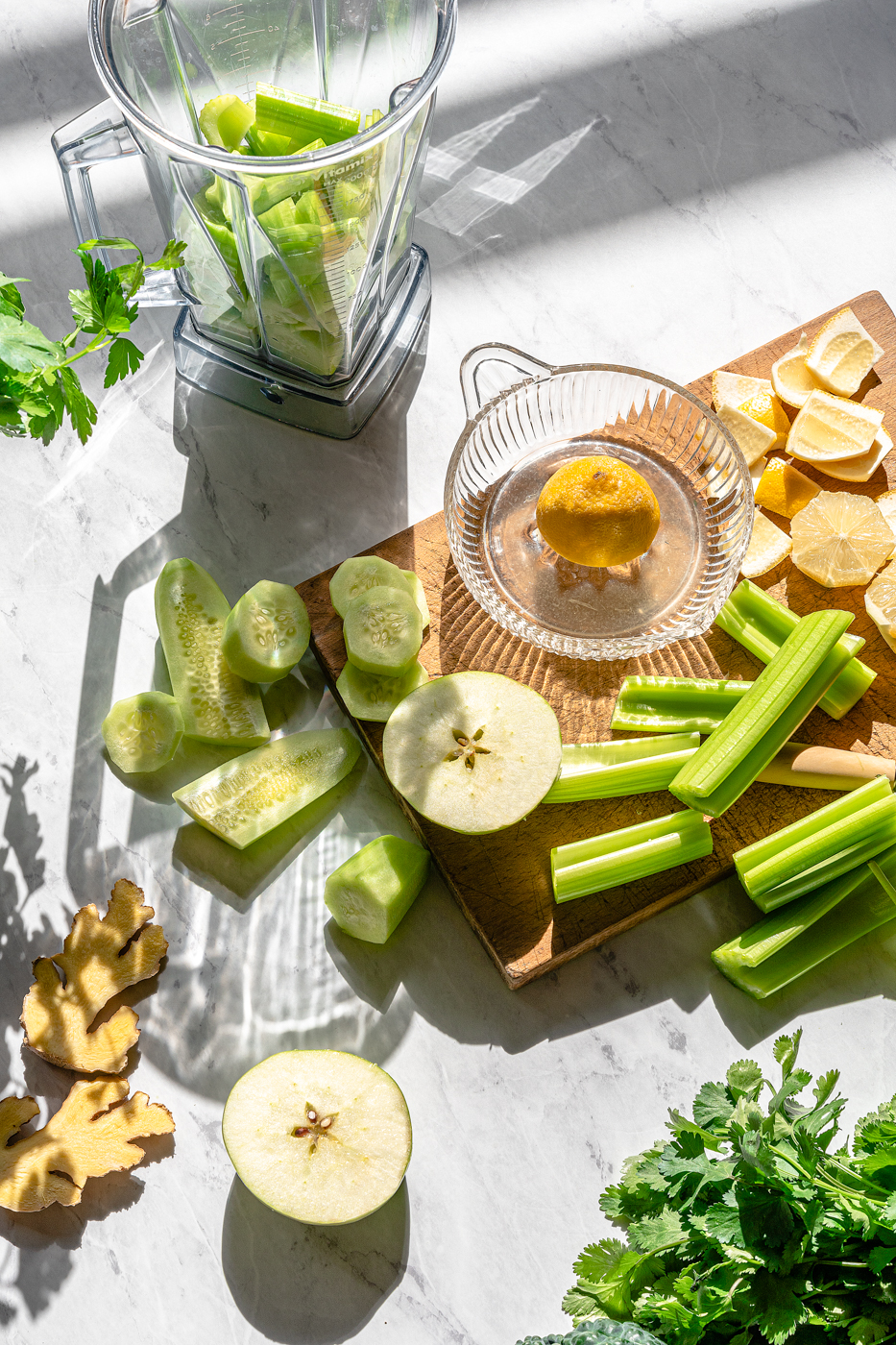 Blender green juice ingredients prepped to go into the blender. Lemon, cucumber, apple, and celery are cut into chunks on a wood cutting board. There is a bunch of celery and parsley in the corner.