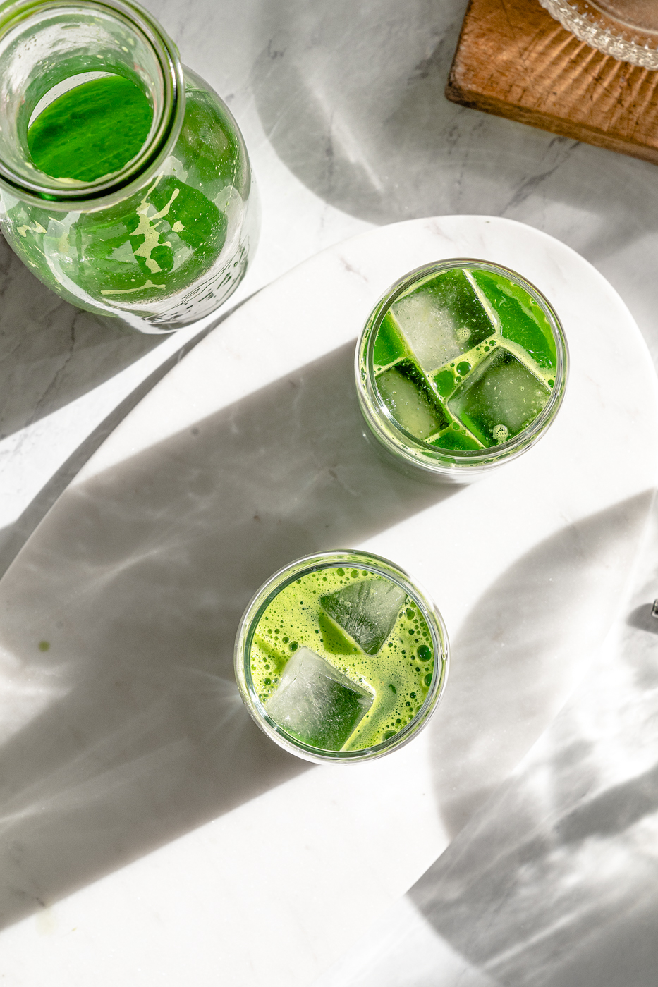 There are two glasses of blender green juice and a jug with extra juice on a marble countertop with long shadows to show drama. 