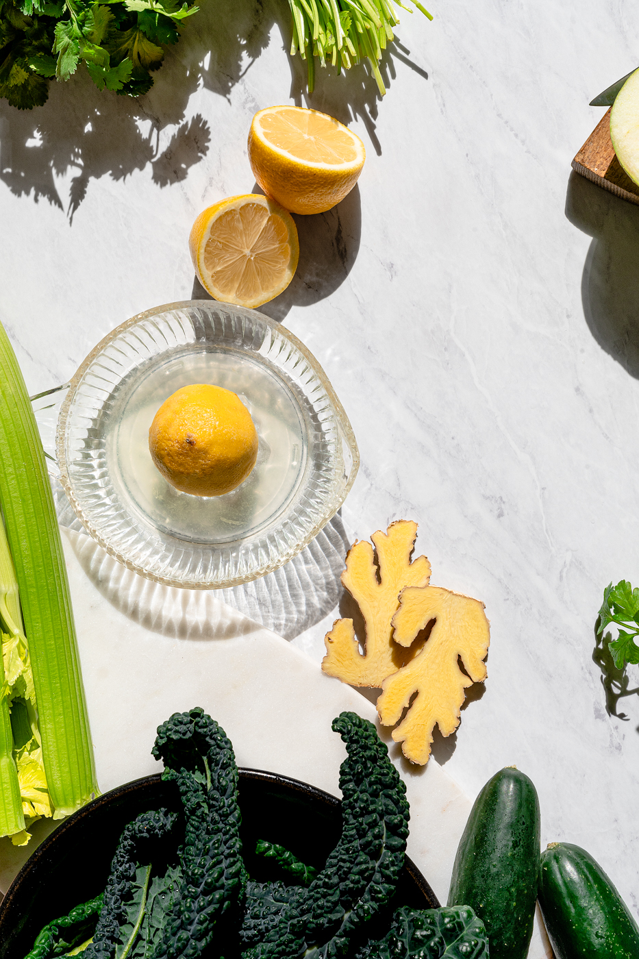 Fresh produce to make blender green juice scattered on a marble counter. There is lemon juice in a juicer, kale, cucumber, celery, ginger, and herbs in the frame.