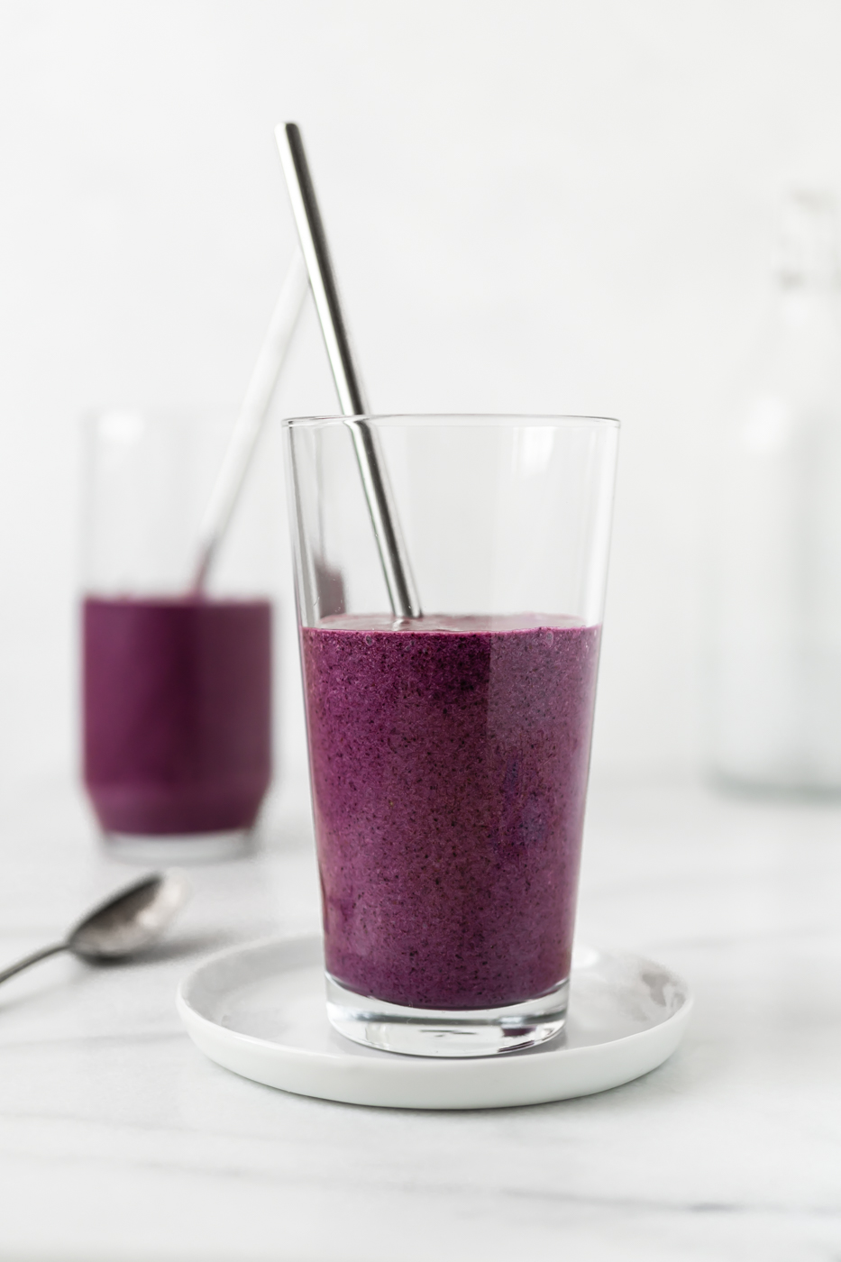 Two purple smoothies in glass cups with metal straws against a white backdrop.