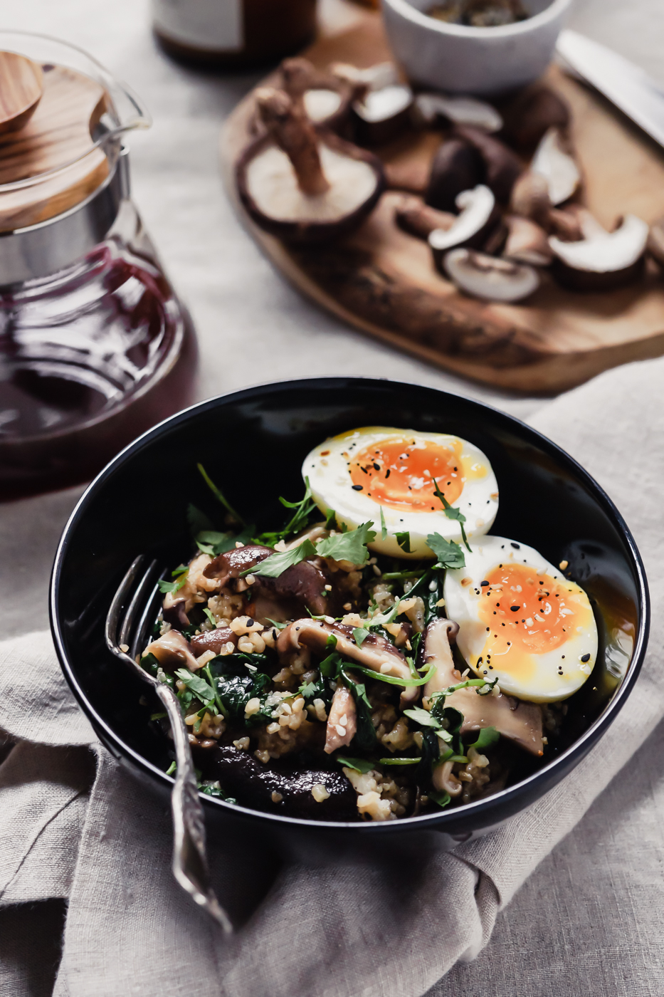 A black bowl that has mushrooms, kale, rice, and a soft boiled egg inside. There is a vessel with coffee in it and a cutting board with mushrooms in the background.