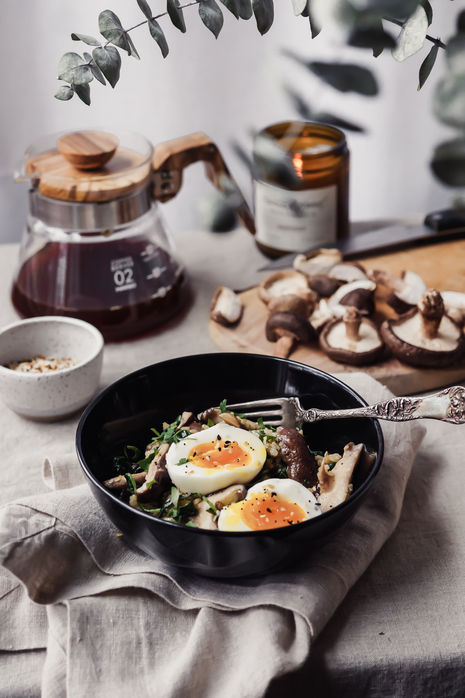 A black bowl that has mushrooms, kale, rice, and a soft boiled egg inside. There is a vessel with coffee in it and a cutting board with mushrooms in the background.