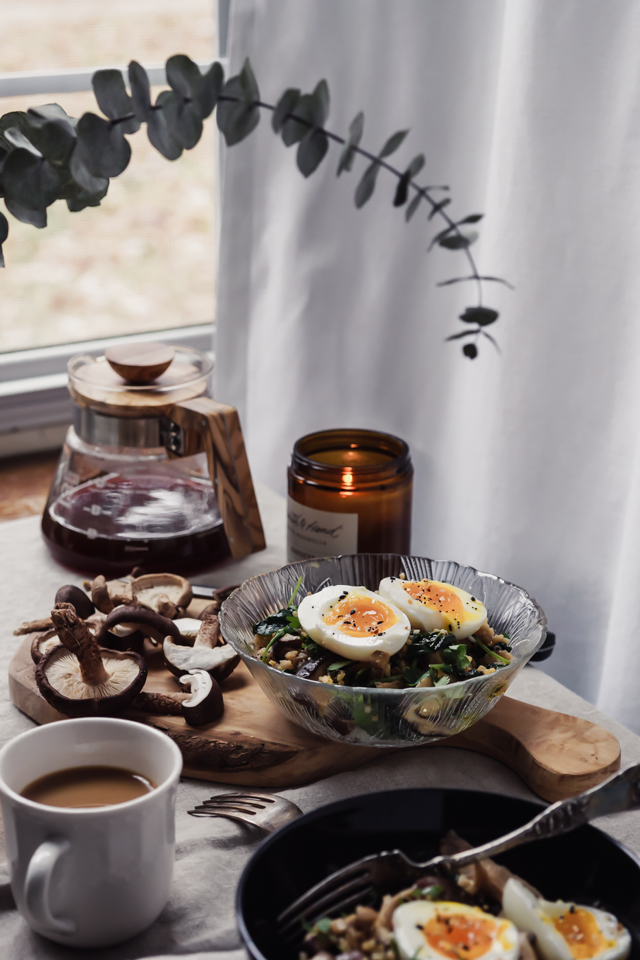 A glass bowl that has mushrooms, kale, rice, and a soft boiled egg inside, set on top of a wood cutting board with mushrooms on top. There is coffee, a candle, and a vase in the photo too.