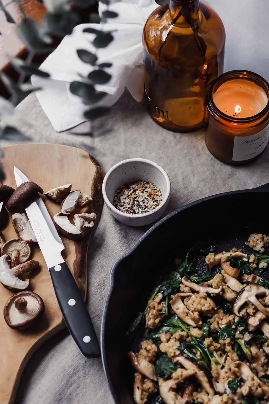 Close up of a wood cutting board with mushrooms and a knife. Everything bagel seasoning, a skillet with vegetables and rice, and a candle are in the picture as well.