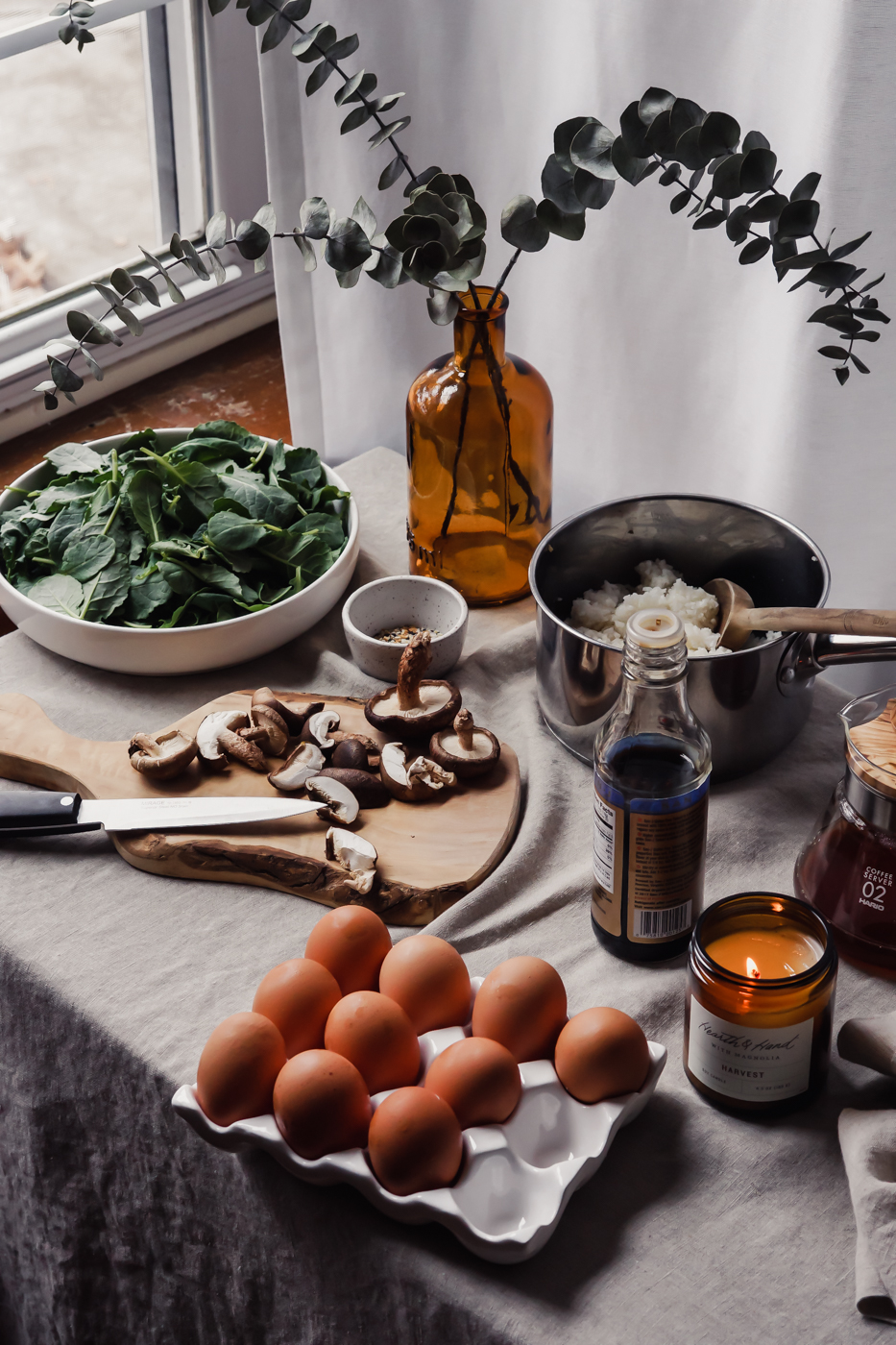Next to a window there is a table that is scattered with ingredients. There are fresh eggs, chopped mushrooms, and a bowl of kale.