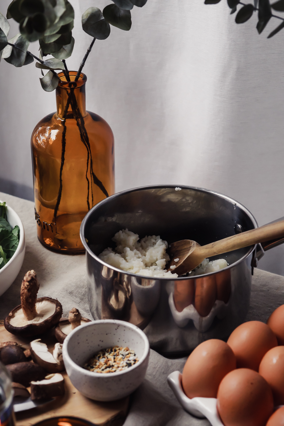 the subject is a pot of rice with a wooden spoon inside. Surrounding the pot are farm fresh eggs, mushrooms, everything bagel seasoning, and a vase.