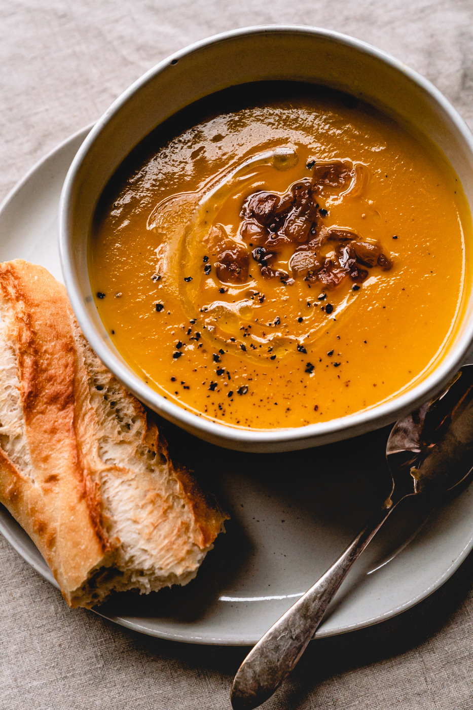 A bowl of creamy Caramelized Onion Butternut Squash Soup on a cream plate with a baguette and silver spoon on the side. 
