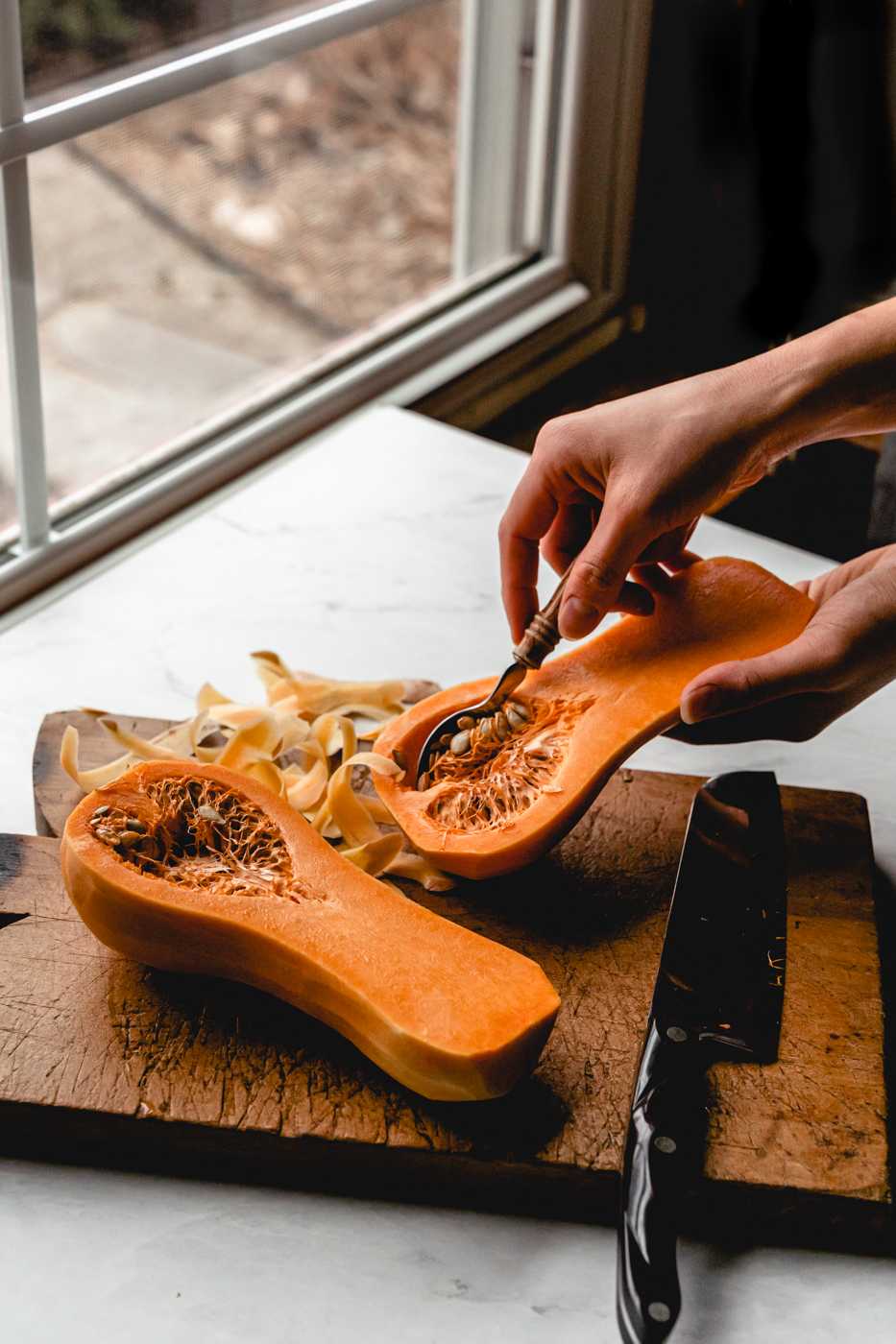 Next to a kitchen window, someone is holding one half of a butternut squash and scooping out the seeds. 