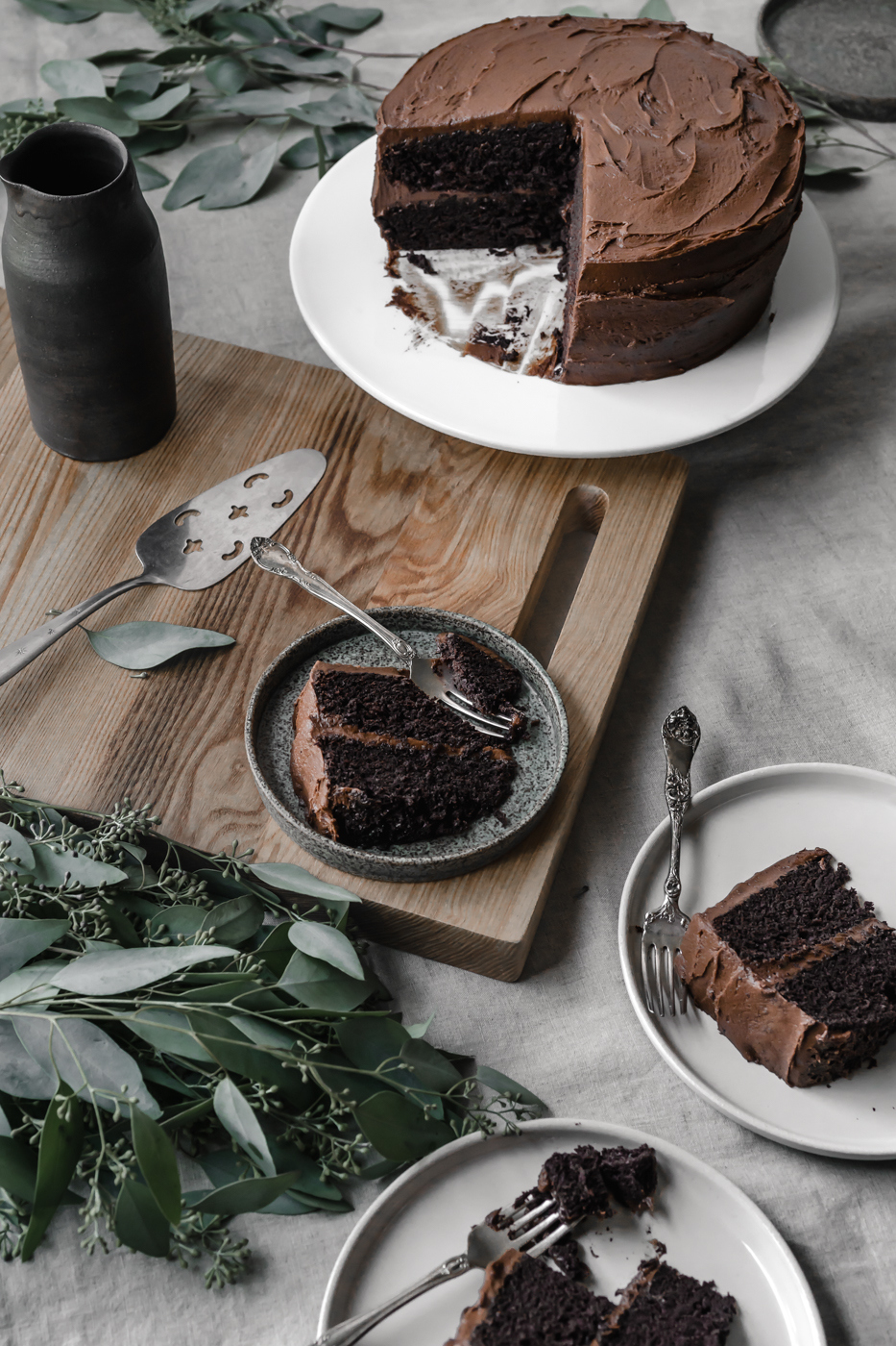 A tablescape photo with the whole two layer chocolate cake sitting on a cake stand with slices of cake surrounding it.