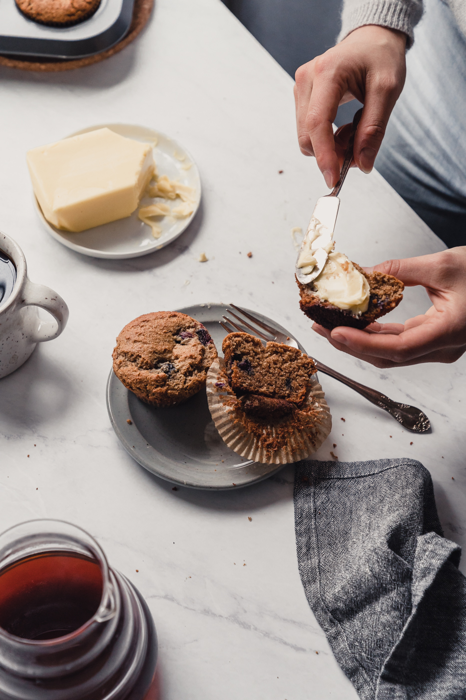 On a marble counter there is a breakfast set up with coffee, butter, a gray linen, and  a plate with two muffins. On the plate, one muffin is whole and one is cut in half and two hands are holding one half of a muffin and spreading butter on it.