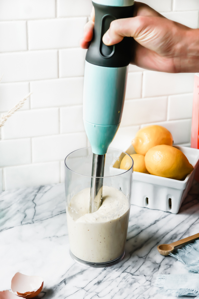hand holding an immersion blender inside a container where a roasted garlic aioli sauce is in the making.