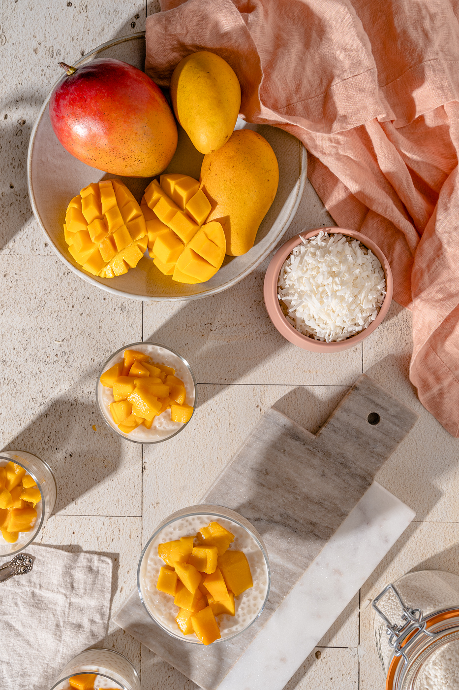 Overhead image of three cups of Mango Sago (coconut milk tapioca pudding) on a stone surface. The shadows are long and the image is bright. There is a bowl of mango, bowl of coconut flakes, and a pink linen at the top of the image.
