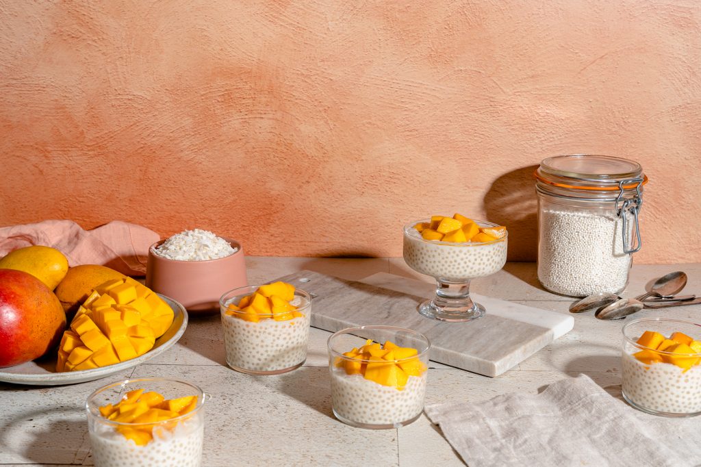 Five small glass cups filled with mango sago (creamy white coconut milk tapioca pudding) on a stone surface with a salmon colored plaster background. Each cup is topped with chunks for juicy mango and the light is glistening off of them. There is a low bowl filled with mangos and a small pink bowl of coconut flakes on the left side of the image.