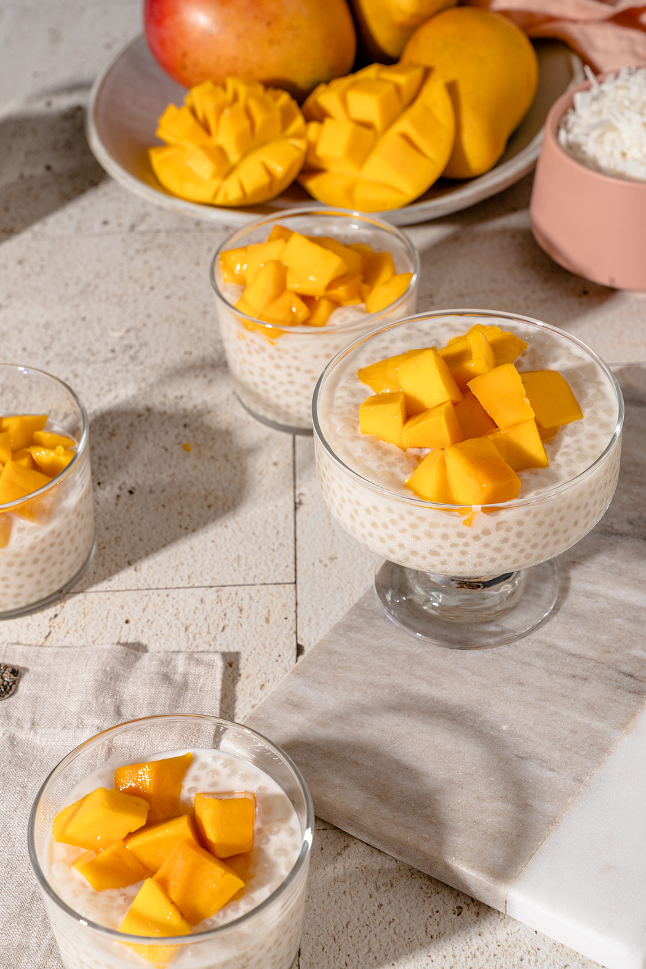 Four small glass cups filled with mango sago (creamy white coconut milk tapioca pudding). Each cup is topped with chunks for juicy mango and the light is glistening off them. There is a low bowl of mangos and a small pink bowl of coconut flakes in the background of the image.