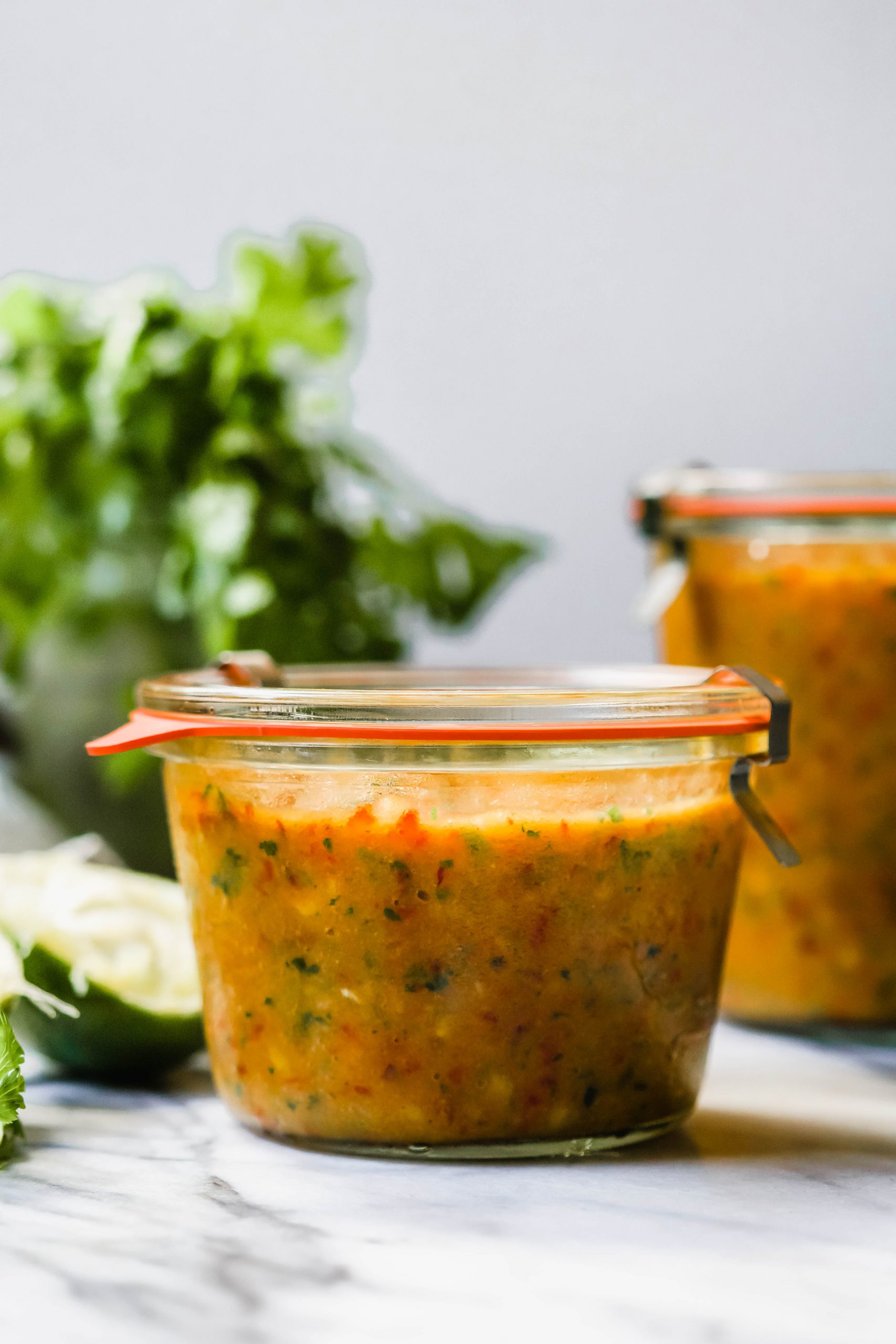 A glass jar of freshly made mango salsa. In the background there is a larger jar, fresh cilantro, and a a squeezed lime.