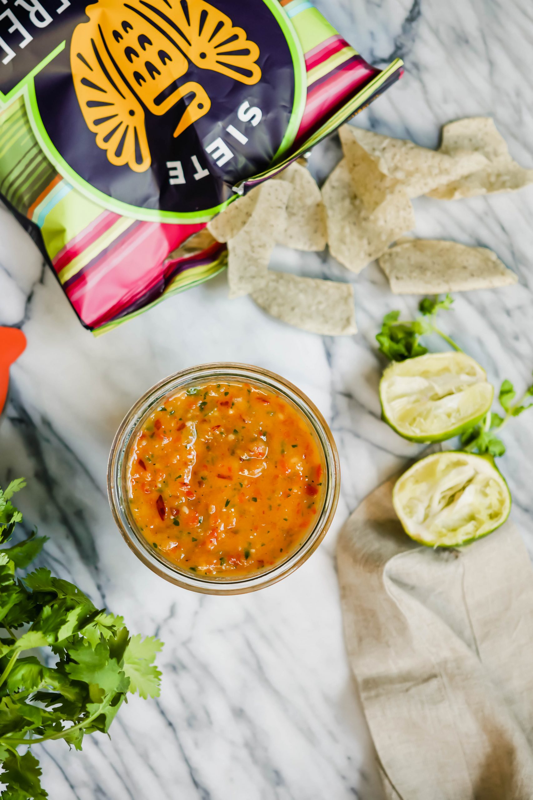 A full jar of freshly made mango salsa with Siete tortilla chips, cilantro, and lime around it.