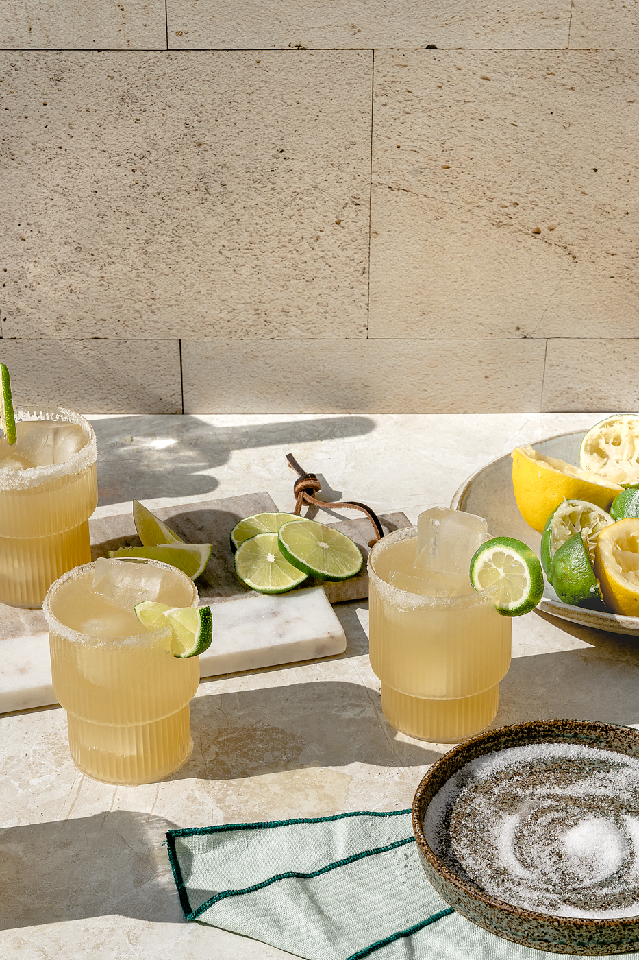 Three ripple glasses with a salt rim are filled with fresh made Classic Margarita and have a lime slice or wedge for a garnish. They are on a stone background and one glass is on a marble cheese board with slices of limes next to the glass. There is a small plate of salt in the bottom right hand corner. There is a low bowl of juiced lemons and limes on the right hand side of the image.