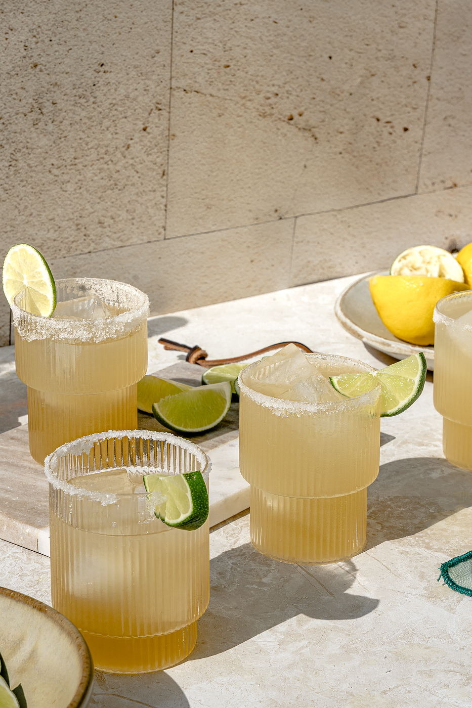 Four ripple glasses with a salt rim are filled with a fresh made Classic Margarita and have a lime slice or wedge for a garnish. They are all at varying heights and against a stone background. 