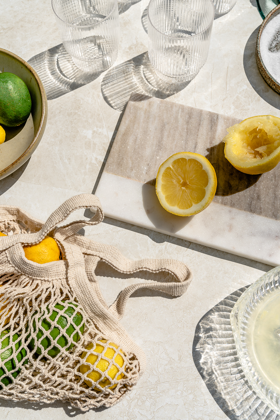 Citrus is prepped for to make a fresh Classic Margarita. On a marble cutting board there is a lemon sliced in half. Lemons and limes are in mesh bag in the bottom left hand side of the image. In the bottom right hand corner, there is an antique citrus juicer filled with lemon just. Three empty ripple glasses are at the top of the image. 