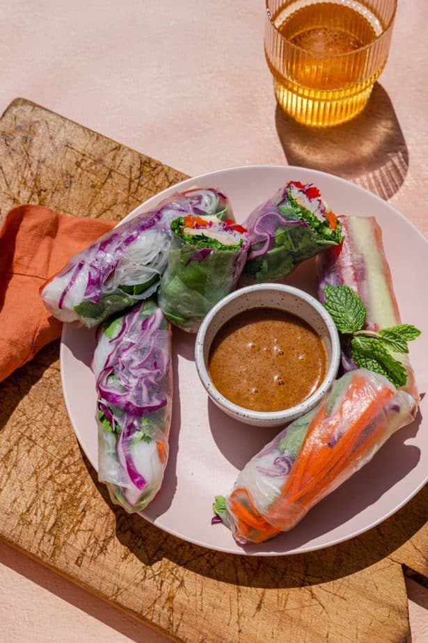 Fresh Vietnamese Spring Rolls with Peanut sauce on a blush pink plate. The plate is sitting on a wooden cutting board with a glass of bear near by. 
