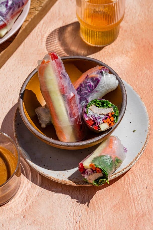 Three Fresh Vietnamese Spring Rolls in a small orange bowl. One of the spring rolls is cut in half so the inside is showing and you can see the fresh vegetables, rice noodles, herbs, and meat. 
