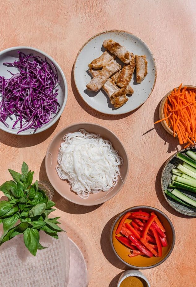 All the filling ingredients spread out in separate dishes. There is thinly sliced red pepper, cucumber, carrot, and cabbage. Cooked pork loin, rice noodles, basil, and rice paper sheets are out too. 