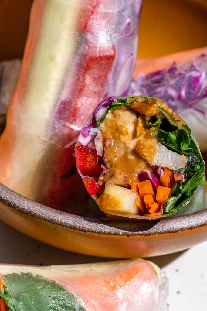 A close up image of  a fresh vietnamese spring roll that is cut in half and has peanut sauce dripping down it. 